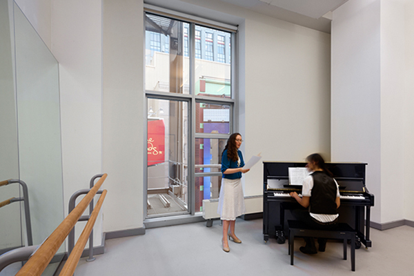 A singing woman stands next to a man playing piano during a music rehearsal in Studio 9C.