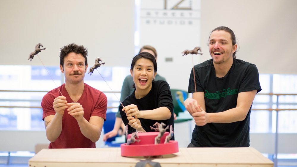 Three actors operate toy horse rod puppets rehearsing a scene.