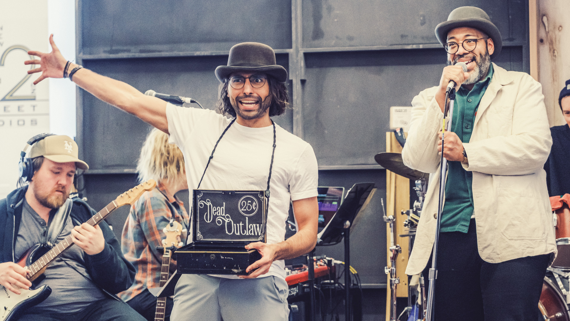 Person in bowler hat with cigar box on string around neck and arm outstretched in front of band and next to singer